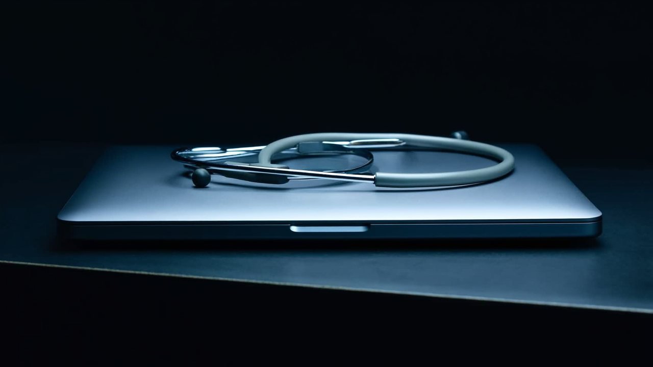 A silver stethoscope resting on a closed laptop on a dark surface, lit by cool ice-blue light against a near-black studio background.